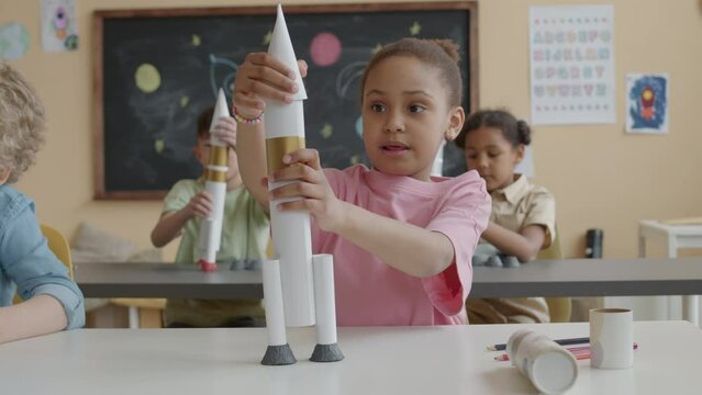 Little African American girl sitting at desk in primary school classroom, building paper rocket and raising it above head while playing during lesson