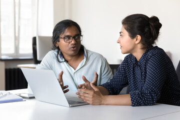 Two serious Indian diverse employees talking at laptop, sitting at workplace, collaborating on project. Corporate mentor explaining work tasks to interns. Developer presenting software to customer