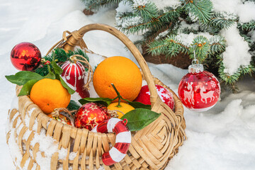 A basket with Christmas decorations stands on the snow outdoors. A basket standing on the snow contains Christmas tree decorations and tangerines. Christmas is coming soon