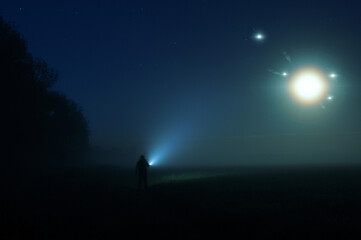 A man with a torch standing in the countryside on a misty summer night. With glowing UFO lights...