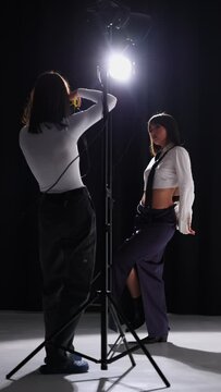 girl photographer taking pictures of asian model in white shirt and tie posing under flashes in studio on black background. vertical video
