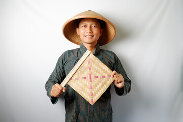 Indonesian farmer wearing Lurik and Caping smiling happy while carrying a bamboo fan
