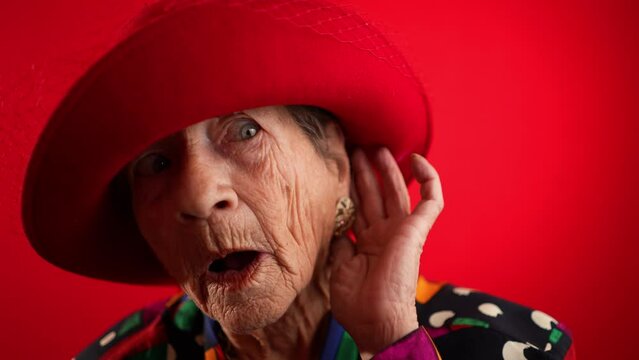 Funny slow motion view of smiling happy crazy elderly woman with no teeth, puts hand to ear to listen to a secret wearing red hat isolated on red background studio
