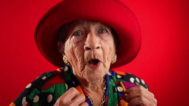 Saying WOW, a happy fisheye closeup portrait caricature of funny elderly woman with red hat isolated on red background in slow motion.