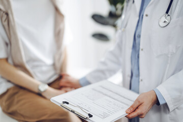 Doctor and patient discussing health exam results. Friendly physician reassuring a young woman by one hand while keeping clipboard with medical papers in another. Medicine concept