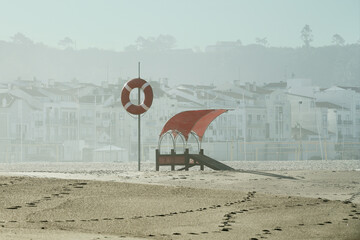 Czerwone pomarańczowe koło ratunkowe na maszcie obok stanowiska ratownika. Mgła, plaża, słońca, lato.