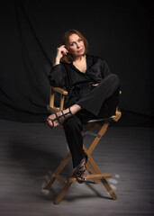 Beautiful adult woman sits on a stylish wooden director's chair on the black background. fashion model in black jacket.