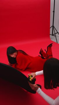 girl photographer takes pictures of an Asian model lying on the floor on her stomach on a red background in a studio in a red sweater and pantyhose. geisha in action in red. vertical video