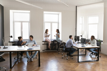 Diverse busy employees working on modern office room, sitting at workplaces, using desktops, talking, discussing project tasks. Contemporary rental office co-working space