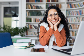 Portrait of happy smiling Indian female teacher sitting at desk in classroom in front of notebook and looking at camera, conducting online classes, young successful tutor, online student.