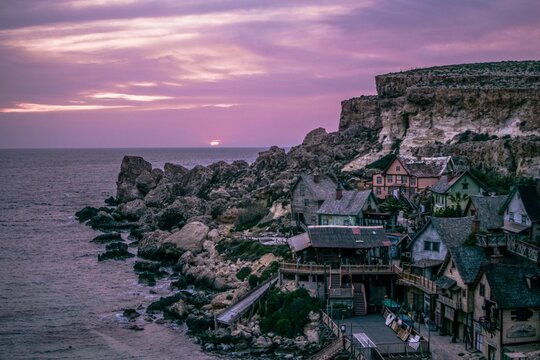 Landscape scene of Popeye Village houses by the sea with purple sunset sky in Mellieha, Malta