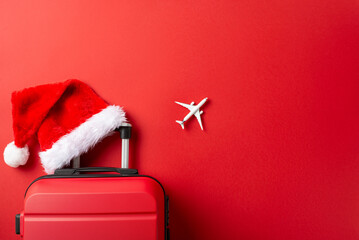 Pack Your Dreams for New Year: Top-view image of a suitcase, miniature airplane model, and Santa's hat on a vibrant red backdrop, offering space for your New Year aspirations - obrazy, fototapety, plakaty