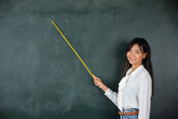 Back to school concept. Asian female teacher smiling with wooden stick pointing to blackboard at school in classroom, Happy beautiful young woman standing hold pointer to back board