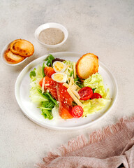 caesar salad with green lettuce, eggs, salmon, cherry tomatoes and croutons on a white ceramic plate, next to a bowl of sauce and a small plate of croutons