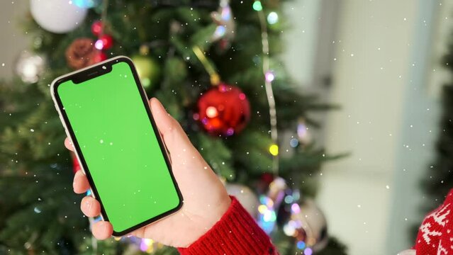 Blonde girl in red sweater and hat looks at green screen of smartphone close-up against background of Christmas tree decorated with garlands and colorful balls. Holiday background.