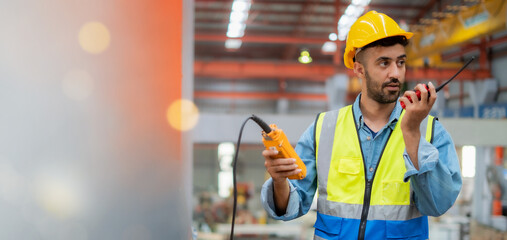 A man in a yellow vest and a hard hat is talking on a walkie talkie. He is in a factory setting