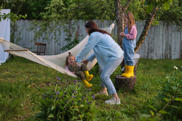 Naklejka na ściany i meble Joyful family moment in nature: Mother playfully interacting with her daughter in a hammock while another child observes, surrounded by a lush garden at sunset. Authenticity and connection