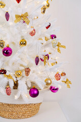 White Christmas tree in a wicker basket decorated shiny and gold decorations, bows, balls, toys and with luminous garland on light background. Merry Christmas and Happy Holidays!