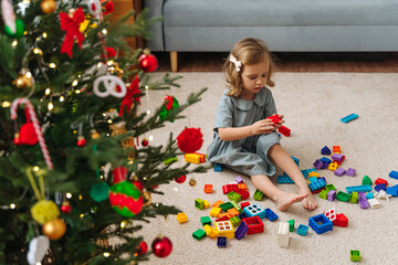 Fototapeta na wymiar A little girl playing with colorful rainbow colored constructor pieces next to the christmas tree. Merry Christmas and Happy Holidays! Christmas gifts for children.