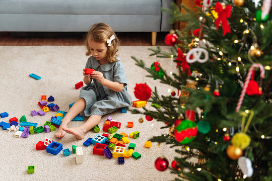A little girl playing with colorful rainbow colored constructor pieces next to the christmas tree. Merry Christmas and Happy Holidays! Christmas gifts for children.