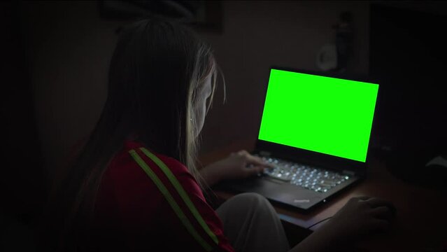 School-age girl sits alone in dark room in front of laptop screen and carefully looks at green screen. Free space for insertion.