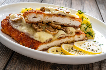 Crispy breaded seared chicken cutlet with fried white mushrooms, cheese and boiled potatoes on wooden table
