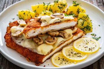 Crispy breaded seared chicken cutlet with fried white mushrooms, cheese and boiled potatoes on wooden table
