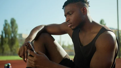 Foto op Plexiglas African American man guy sportsman scrolling smartphone sport and technology in city male use mobile phone having break outdoors on sports court athlete using cellphone outside relaxing post-workout © Yuliia