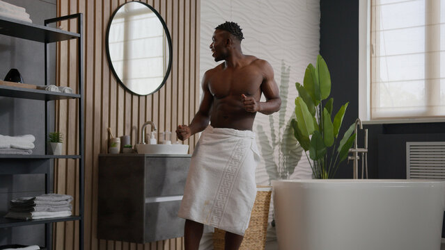 Funny happy ethnic man African American male muscular athletic guy good physical shape dancing with bath towel on hips morning energy active dance to music in home bathroom after washing in bathtub