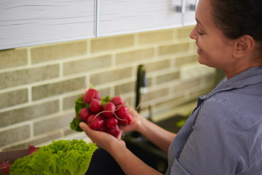 Overhead View Woman Holding Fresh Radish, Standing By Kitchen Counter With A Cardboard Box Full Of Delivered Vegetables