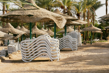 Empty beach with palm, white sunbeds, wicker umbrellas waiting for new summer holiday season tourists. Abandoned equipment. Inspirational tourism travel. Stack of sun loungers. End of swimming season.