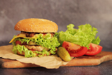 Double cheeseburger and vegetables on a gray background. Juicy tasty burger close-up on a wooden cutting board, side view. Fast food