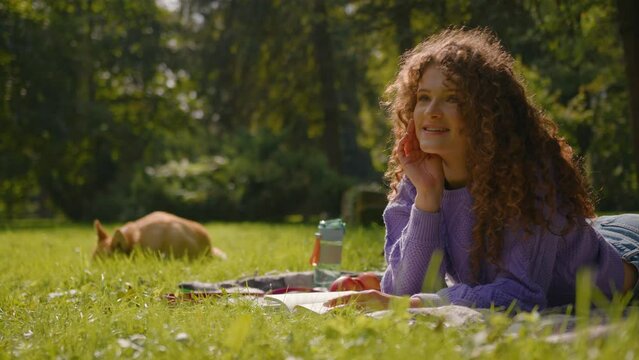 Caucasian happy girl handler pet owner reading book on grass lawn outdoors summer weekend with playful little dog corgi in park nature smiling young woman student read literature relax rest outside
