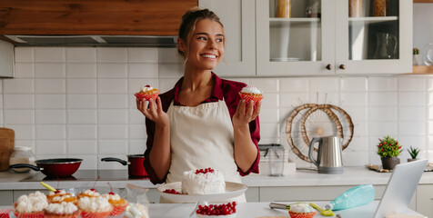 Beautiful female baker holding homemade muffins and smiling while standing at the domestic kitchen