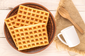 Three freshly baked aromatic waffles on a clay plate with a cup and a spoon on a wooden table, macro, top view.