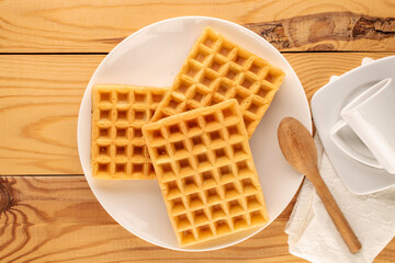 Three freshly baked aromatic waffles on a ceramic white plate with a cup and a spoon on a wooden table, macro, top view.