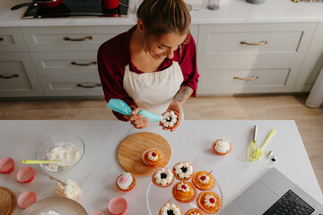 Top view of female confectioner applying whipped cream on cakes while standing at the kitchen