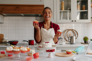 Happy female baker holding homemade muffins while standing at the domestic kitchen