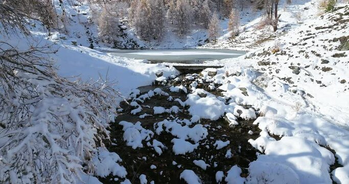 Lac Bleu Arolla lake frost in autumn colors in winter time snow covered Swiss mountains in Switzerland orange colors daylight flowing stream river aerial by drone