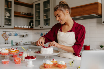 Smiling female confectioner decorating cake with berries while standing at the domestic kitchen