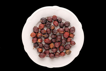 Several dry rosehip berries with a white ceramic plate on a black background, macro, top view.