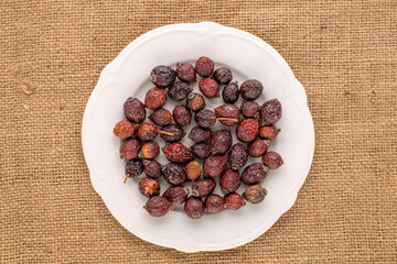Several dry rose hips with white ceramic plate on jute cloth, macro, top view.