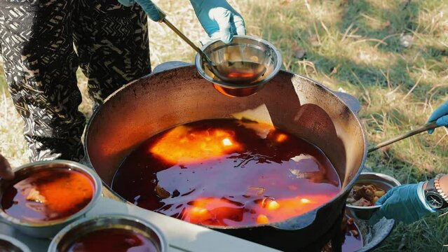 Close-up top view of a huge cauldron of bograch or borscht outdoors, while female hands use a kitchen ladle to scoop soup into metal plates. Camping season