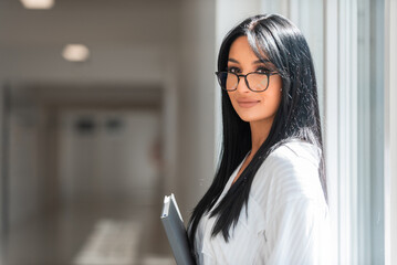 Portrait of a young happy business woman standing in office corridor near window or female college student on the university campus.