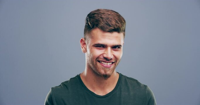 Confident, wink and face of man on gray background for happiness, flirting and romance gesture in studio. Smile, body language and portrait of happy person with reaction for secret, comic and emoji