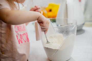 Small toddler girl in the kitchen mixing flour and baking soda with spoon in a measuring cup while...
