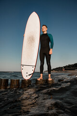 Handsome young male surfer in wetsuit standing on wooden stump holding surfboard and looking away close up