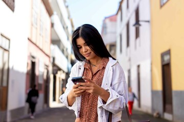 Young Hispanic woman sending message on her mobile phone walking around city