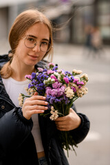 Photo of the girl with a brown hair and glasses, who stands on a city street with a bouquet colorful flowers