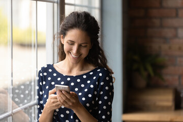 Smiling woman using smartphone, standing near window at home, happy young female looking at phone screen, chatting with boyfriend in social network or shopping online, reading pleasant news